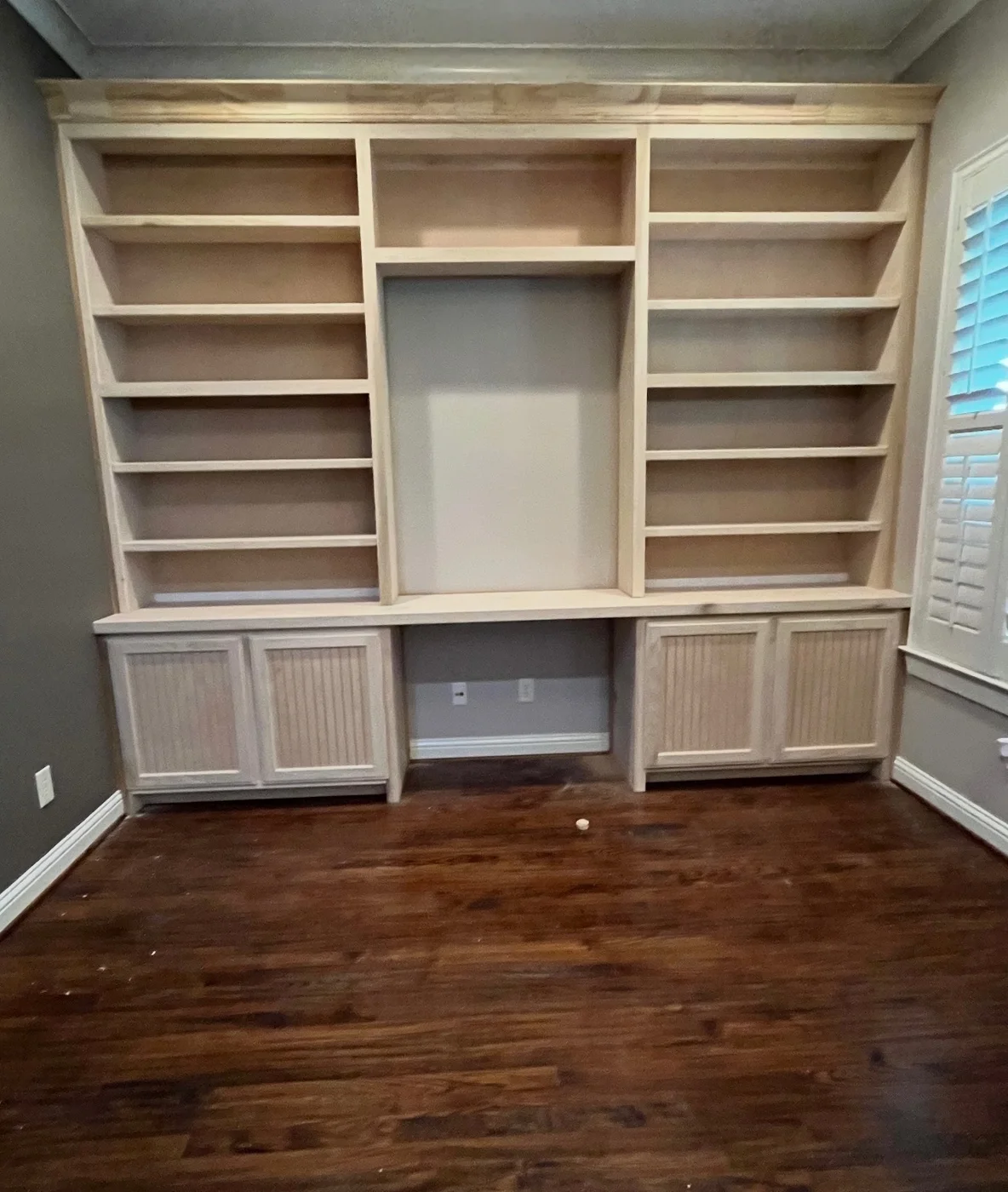 Wall-to-wall unfinished wood shelving with lower cabinets and a centered media opening.